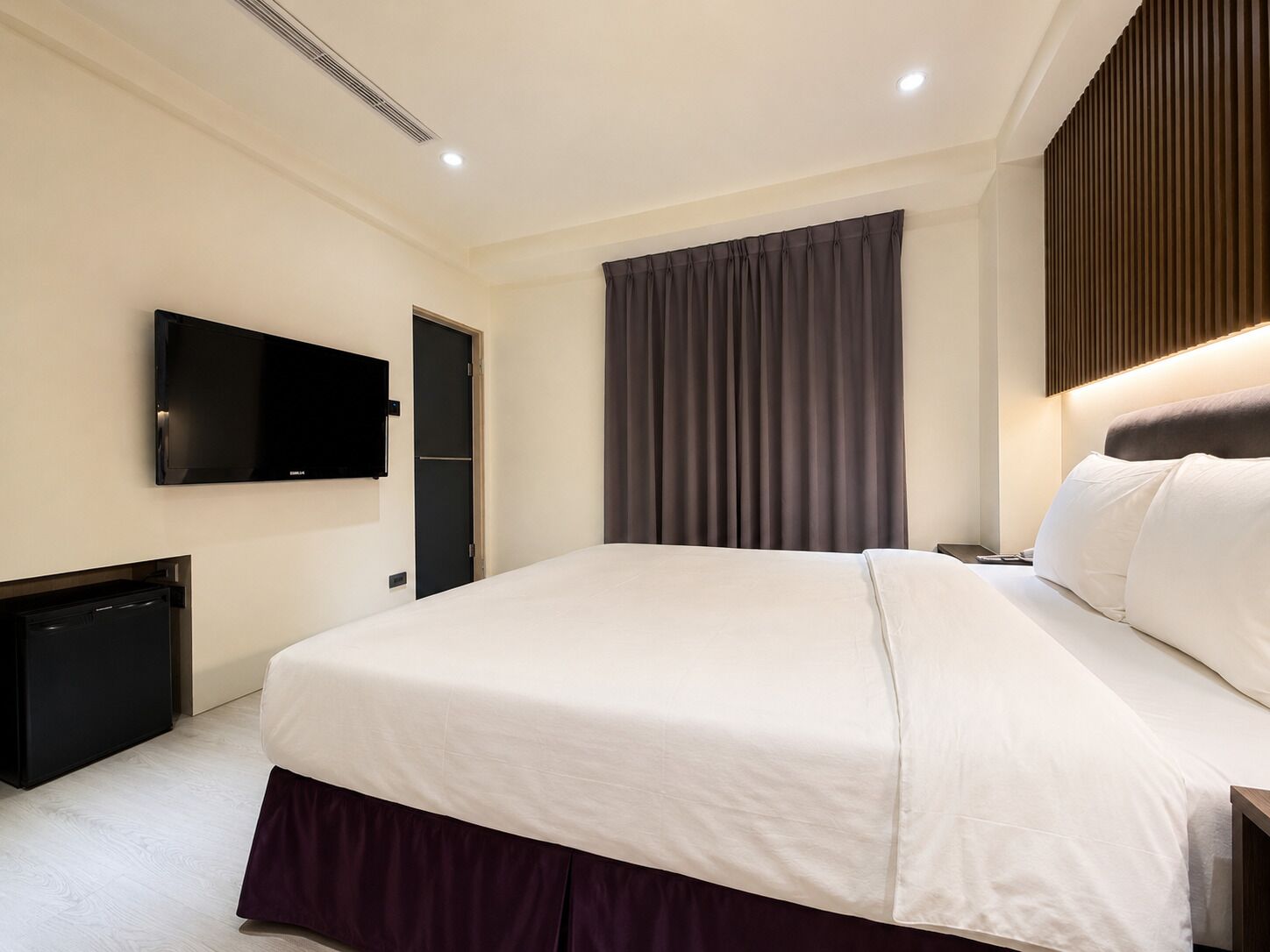 Una habitación de hotel moderna con una cama grande, un televisor de pantalla plana en la pared y un cabecero de color oscuro.