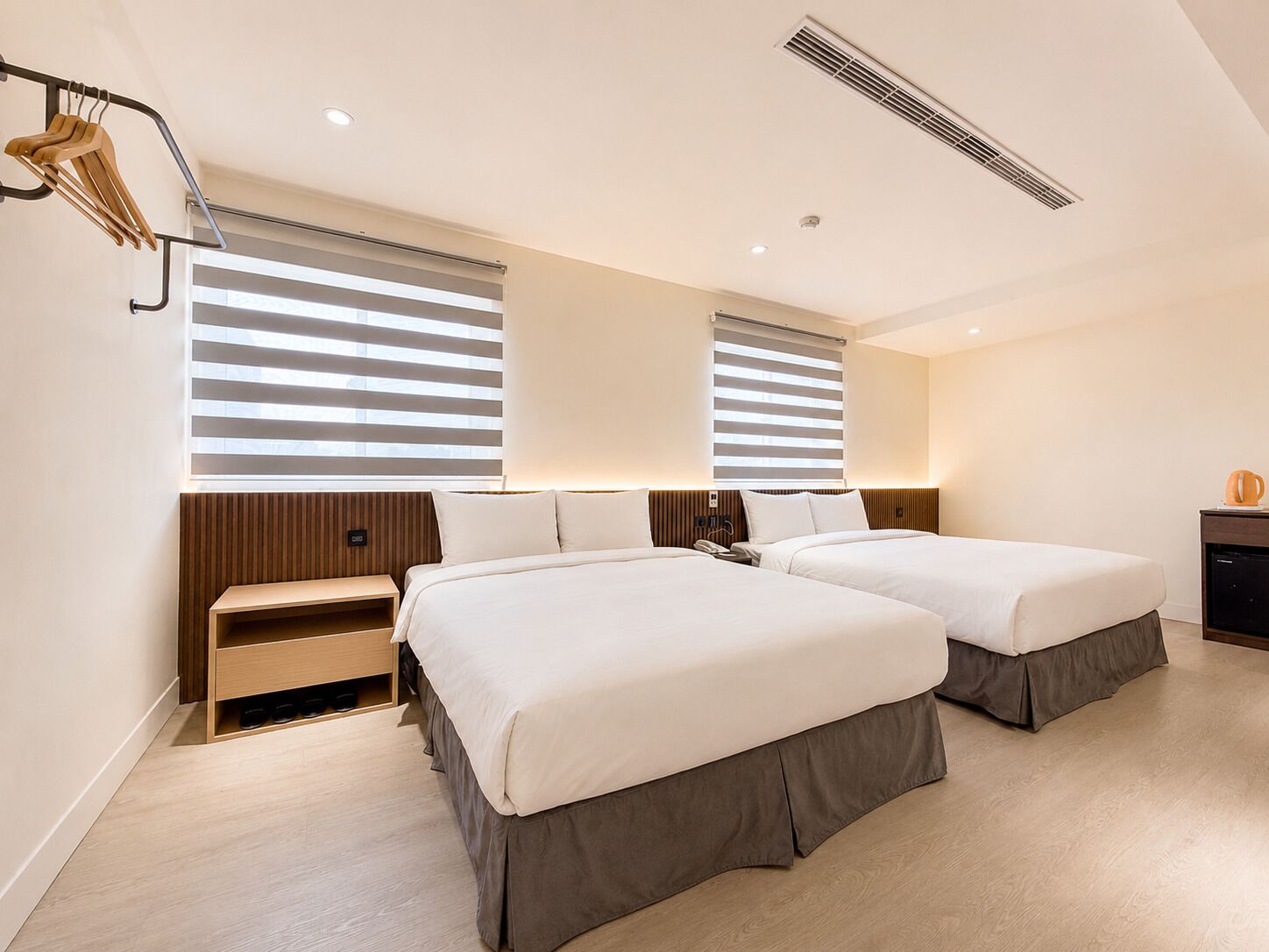 A modern hotel room with two beds, a wooden headboard, a bedside table, and a small cabinet.