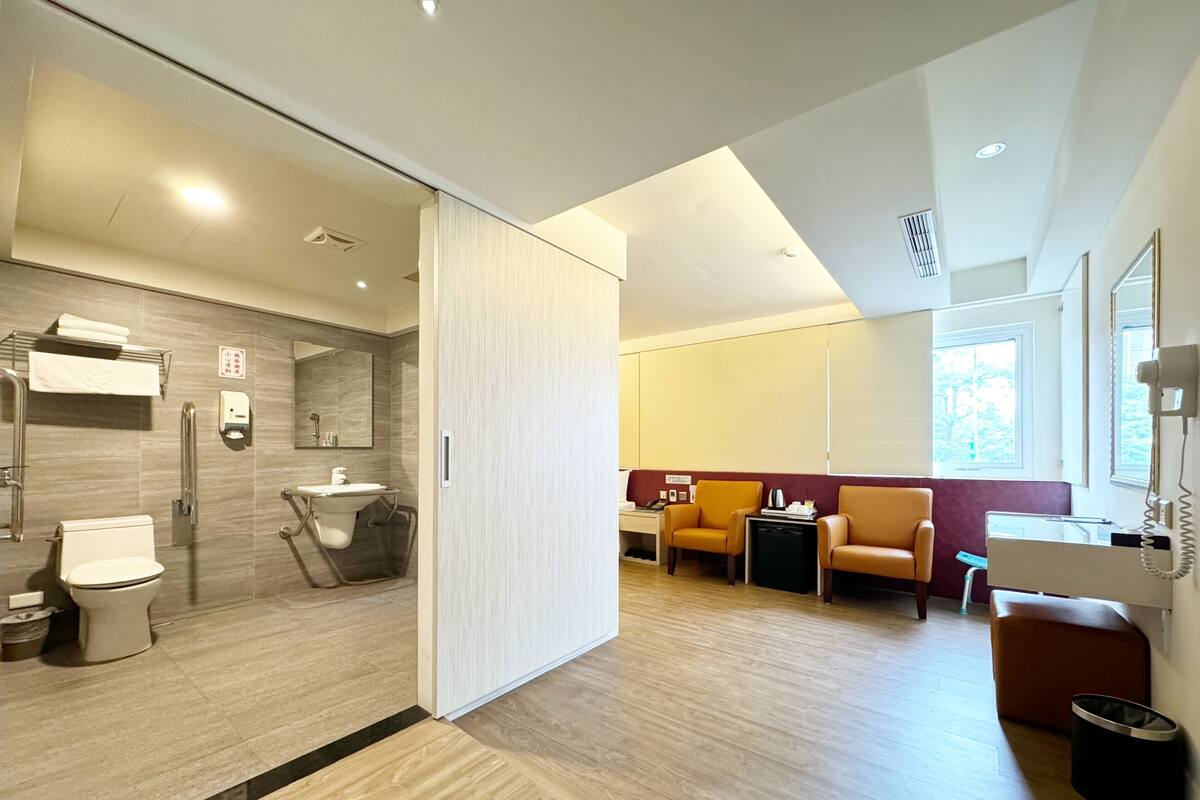 Une chambre d’hôtel moderne dotée d’une salle de bain, d’un coin salon avec deux fauteuils, d’un bureau et d’une grande fenêtre.