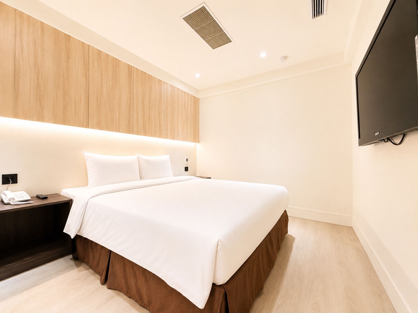 Une chambre d’hôtel équipée d’un lit, d’une table de chevet, d’un téléphone, d’une télécommande, d’un téléviseur fixé au mur et d’une bouche d’aération au plafond.