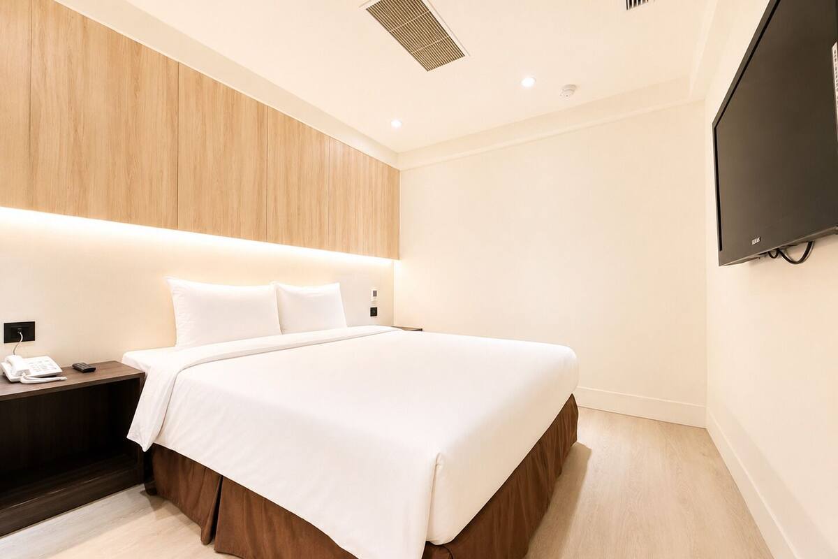 Une chambre d’hôtel équipée d’un lit, d’une table de chevet, d’un téléphone, d’une télécommande, d’un téléviseur fixé au mur et d’une bouche d’aération au plafond.