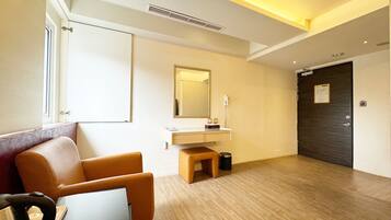 A hotel room with a wooden floor, a desk with a chair, a mirror, a vanity with a mirror, a door, and a window.