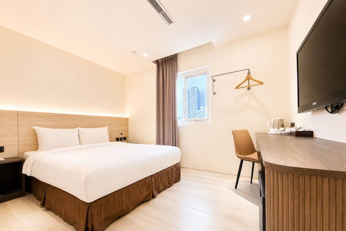 Une chambre d’hôtel comprenant un lit, une table de chevet, une chaise, un bureau, une télévision et une fenêtre avec des rideaux.