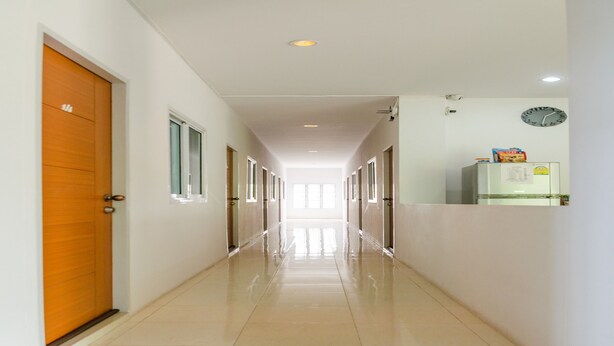 A long, well-lit hallway with a wooden door on the left, a clock on the right, and a refrigerator in the background. - Be My Guest (Ubon Ratchathani)