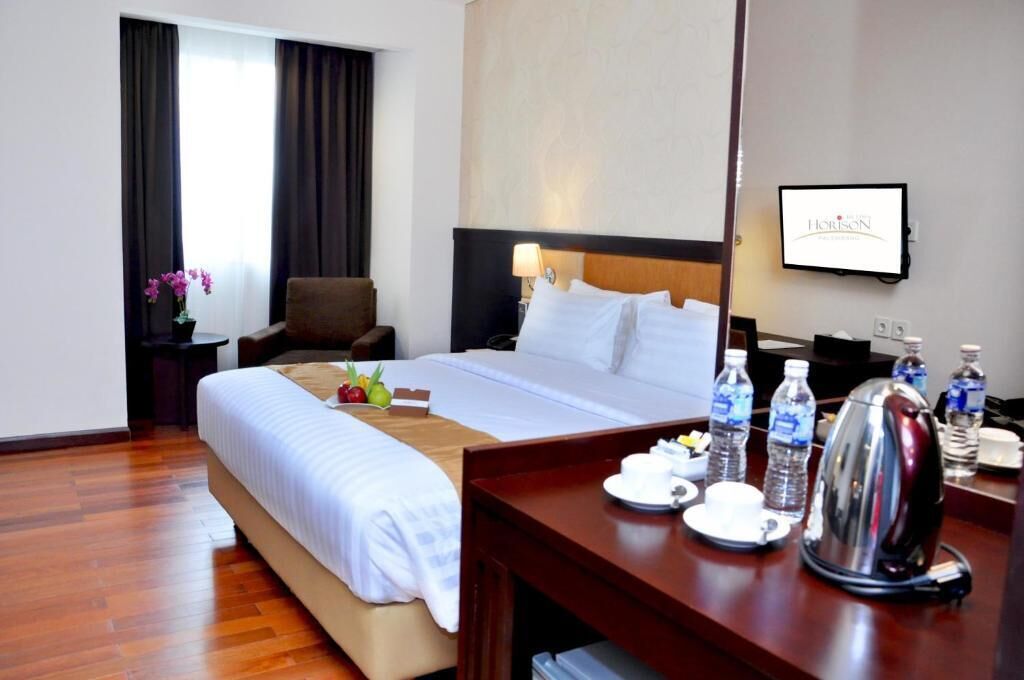 A hotel room with a bed, a desk with a kettle, water bottles, and a TV on the wall.
