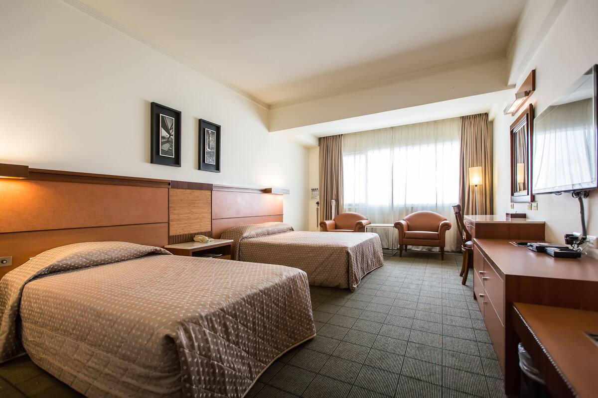 Quarto de hotel com duas camas, uma escrivaninha, uma cadeira, uma televisão e uma janela com cortinas.