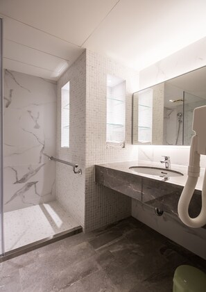 A modern bathroom with a glass shower enclosure, a marble-tiled wall, a sink with a handheld showerhead, and a large mirror.