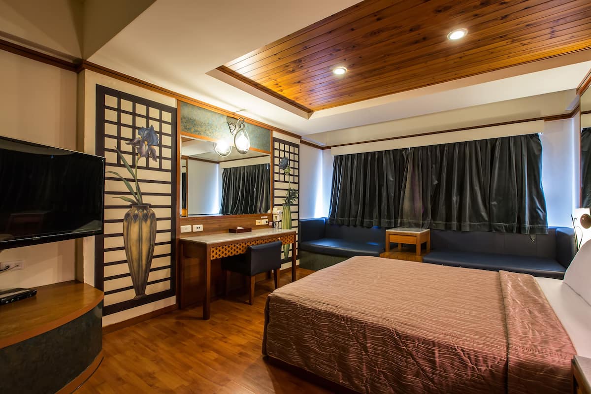 Quarto de hotel com teto de madeira, uma cama grande, uma mesa de trabalho com cadeira, televisão e uma janela ampla com cortinas.