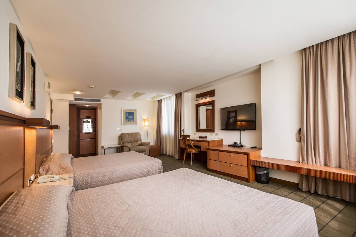 Quarto de hotel com duas camas, uma escrivaninha, uma cadeira, uma televisão e banheiro.