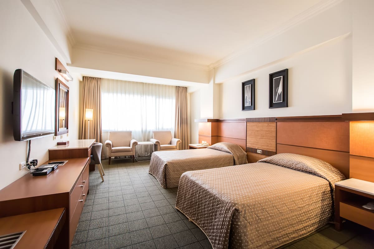 Quarto de hotel com duas camas, uma mesa, uma cadeira, televisão e janela com cortinas.