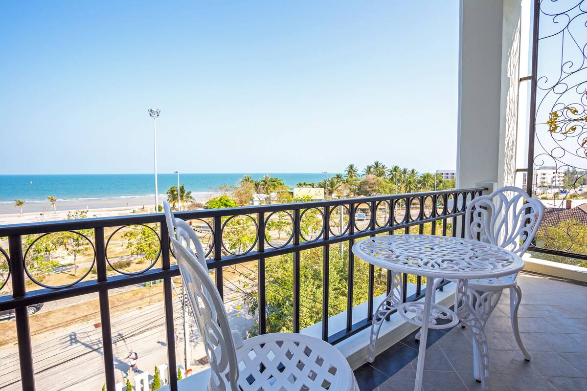 A balcony with white metal chairs and a table, overlooking a beach with palm trees and a clear blue sky.