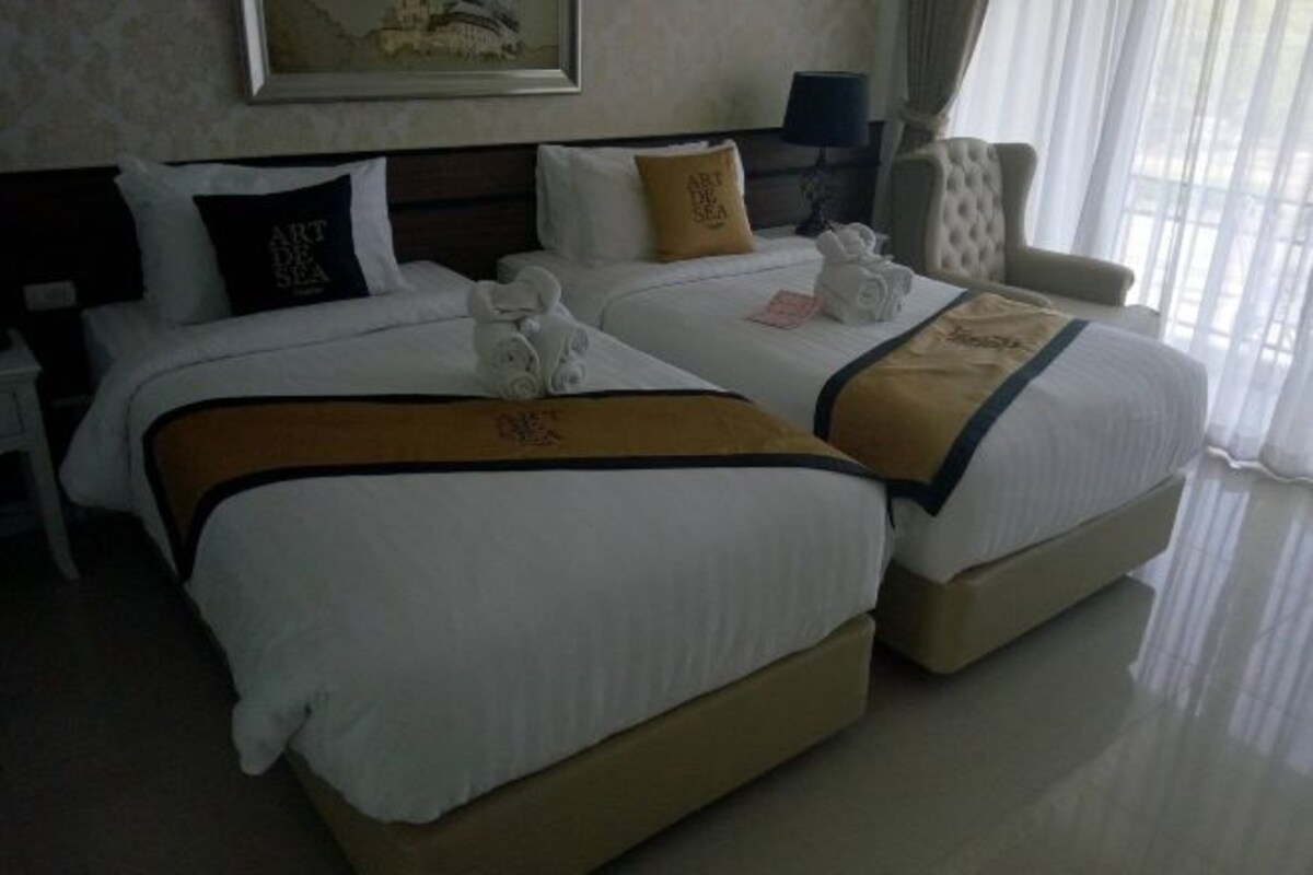 A hotel room with two beds, each with white linens and a brown accent. There are towels on the beds, a lamp on the nightstand, and a chair by the window.