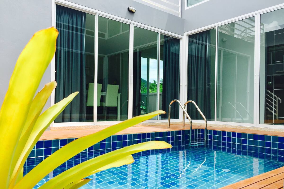 A swimming pool with a wooden deck and blue tiles, surrounded by large windows with curtains.