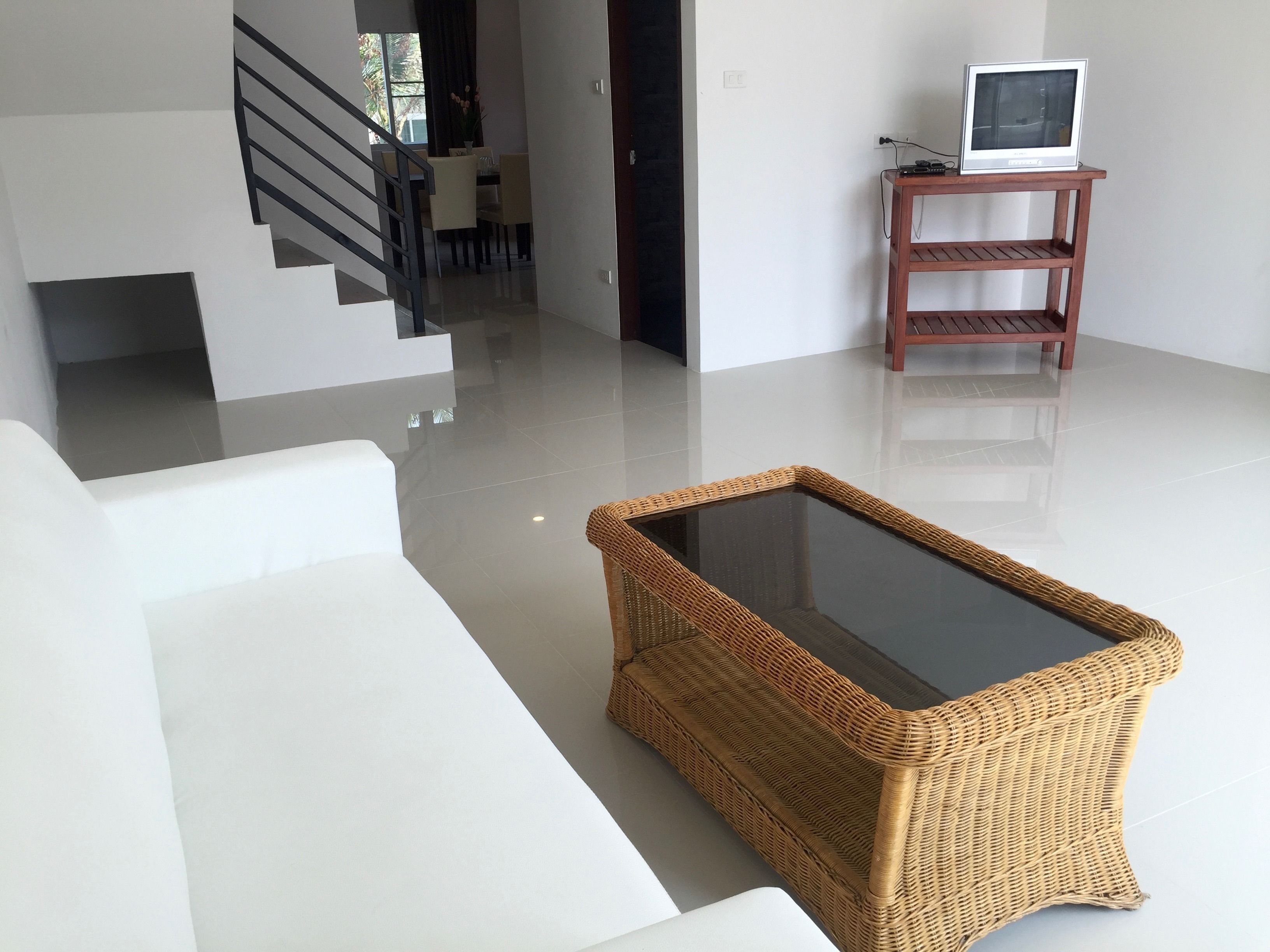 A modern living room with a white sofa, a wicker coffee table with a glass top, a wooden TV stand with a television, and a staircase leading to another room.