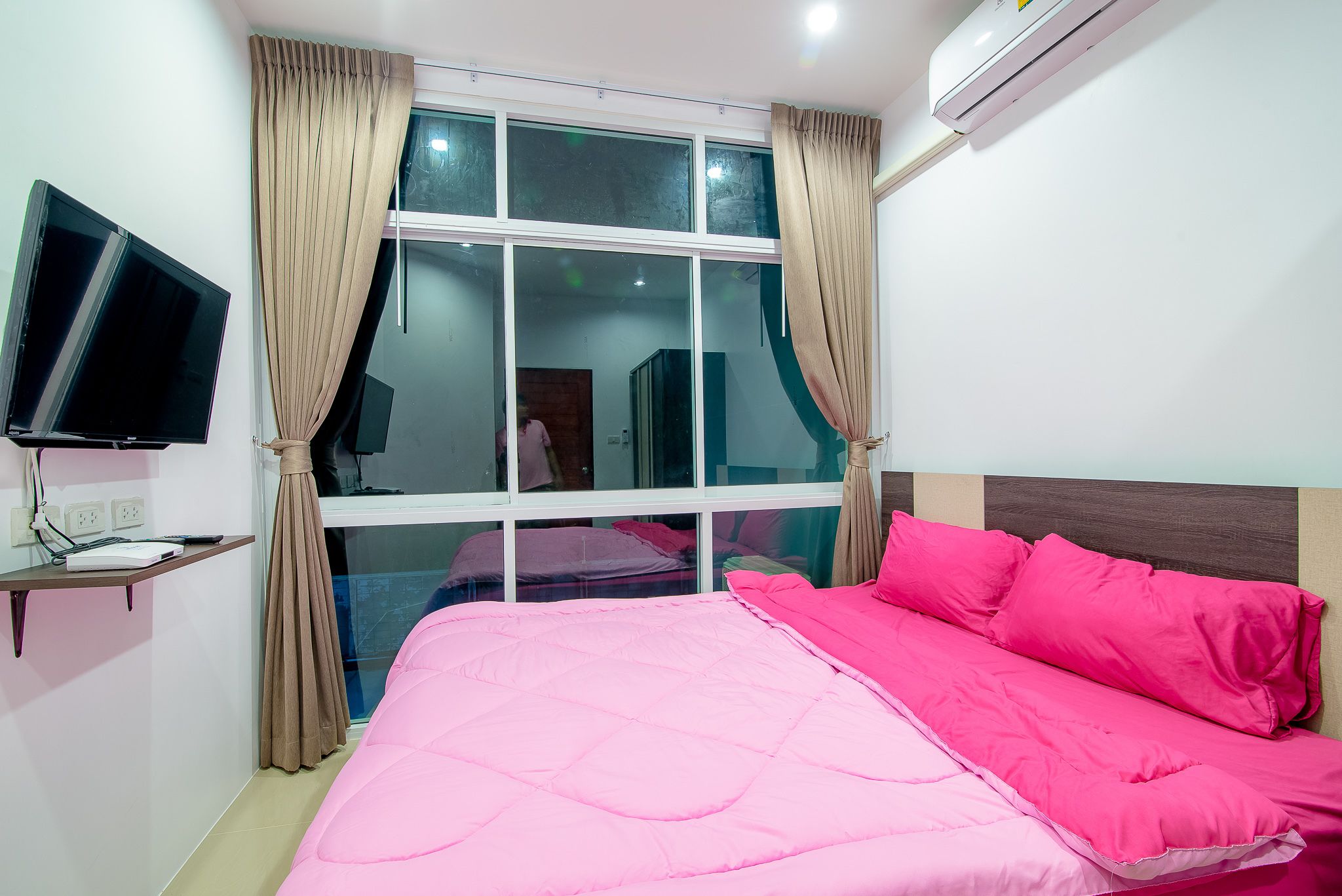 A hotel room with a large bed, a flat-screen TV, a window with curtains, and an air conditioning unit.