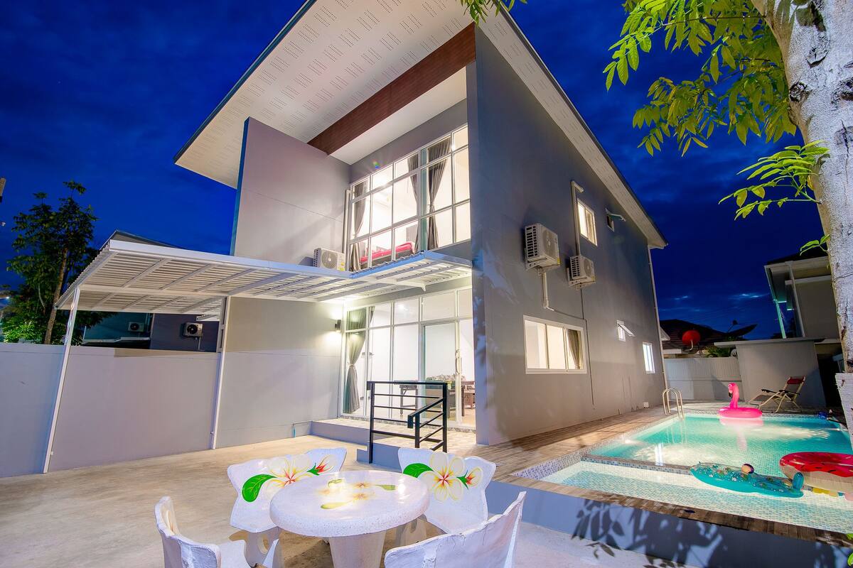 A modern house with a swimming pool, a covered patio, and a dining area with white chairs and a table.