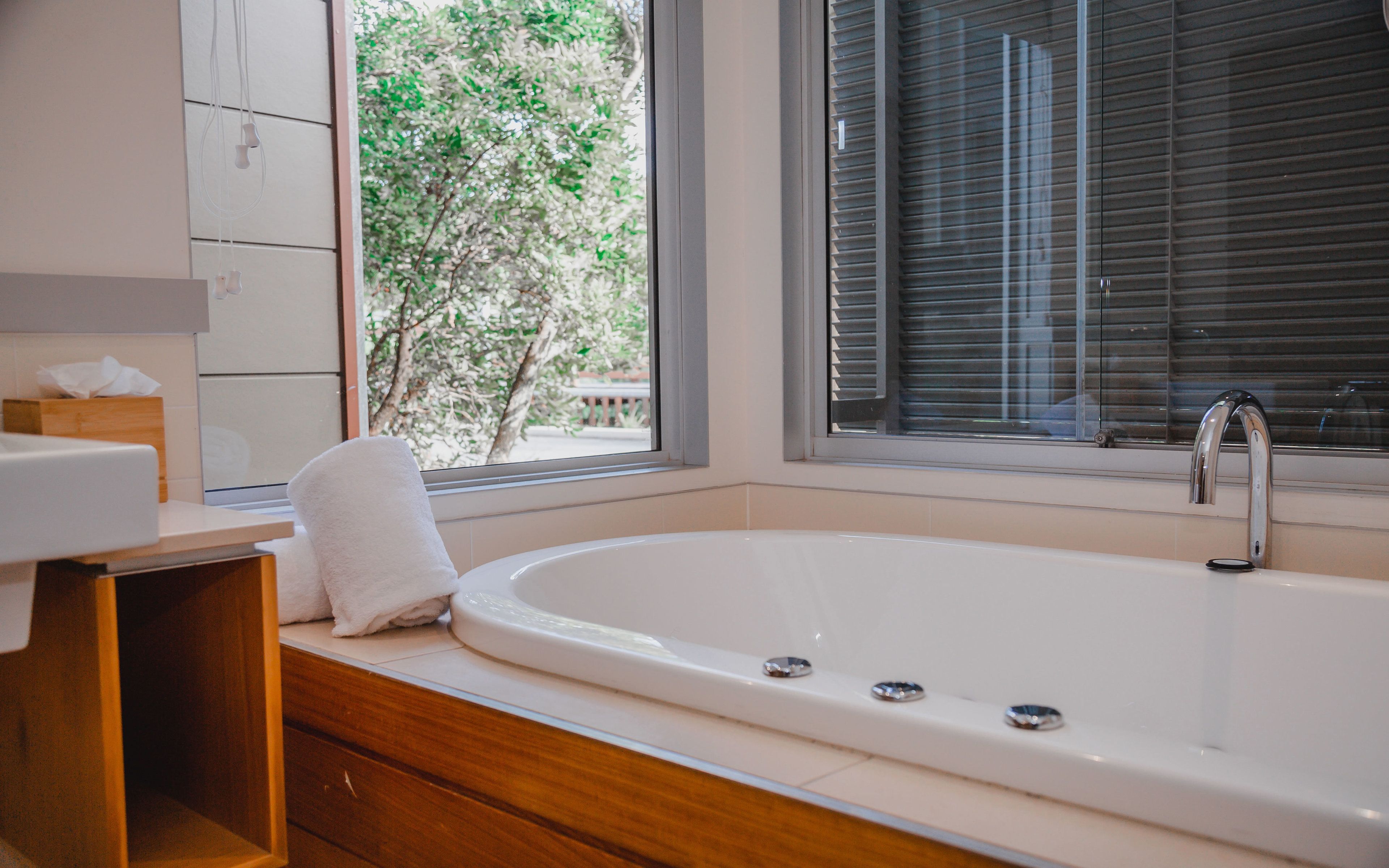 Un baño moderno con una bañera grande, tocador de madera y una ventana con persianas.