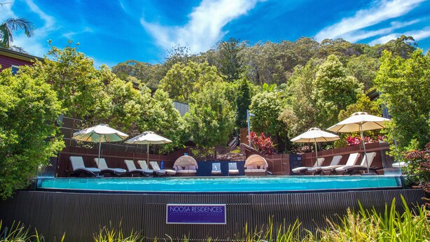 A pool area with lounge chairs and umbrellas, surrounded by lush greenery and a clear blue sky.