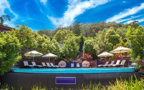 A pool area with lounge chairs and umbrellas, surrounded by lush greenery and a clear blue sky.