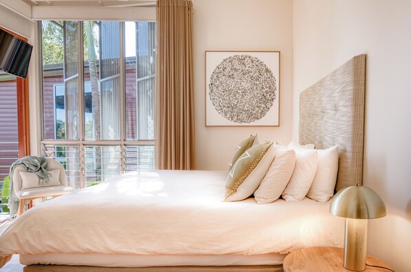 A bedroom with a large bed, a headboard, a bedside table, a lamp, a chair, a window with curtains, and a framed artwork on the wall.