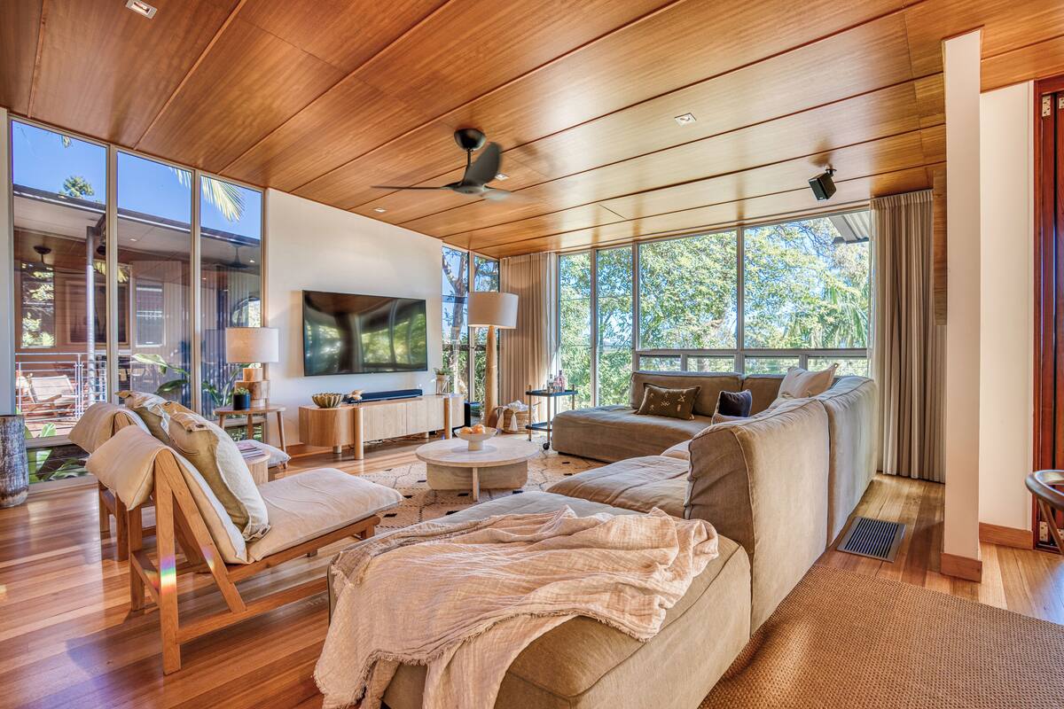 Un salon spacieux doté d’un plafond en bois, de grandes fenêtres, d’un téléviseur à écran plat et d’un coin salon confortable.