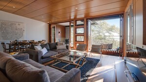 A spacious living room with a large wooden ceiling, a dining area with a table and chairs, and a view of the outdoors through large sliding glass doors.