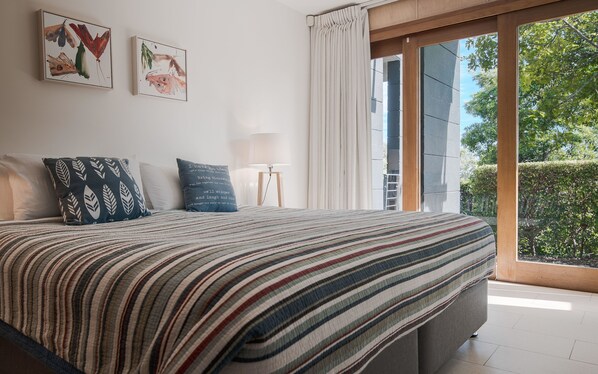 A bedroom with a large bed, two pillows, a bedside table with a lamp, and a window with curtains.
