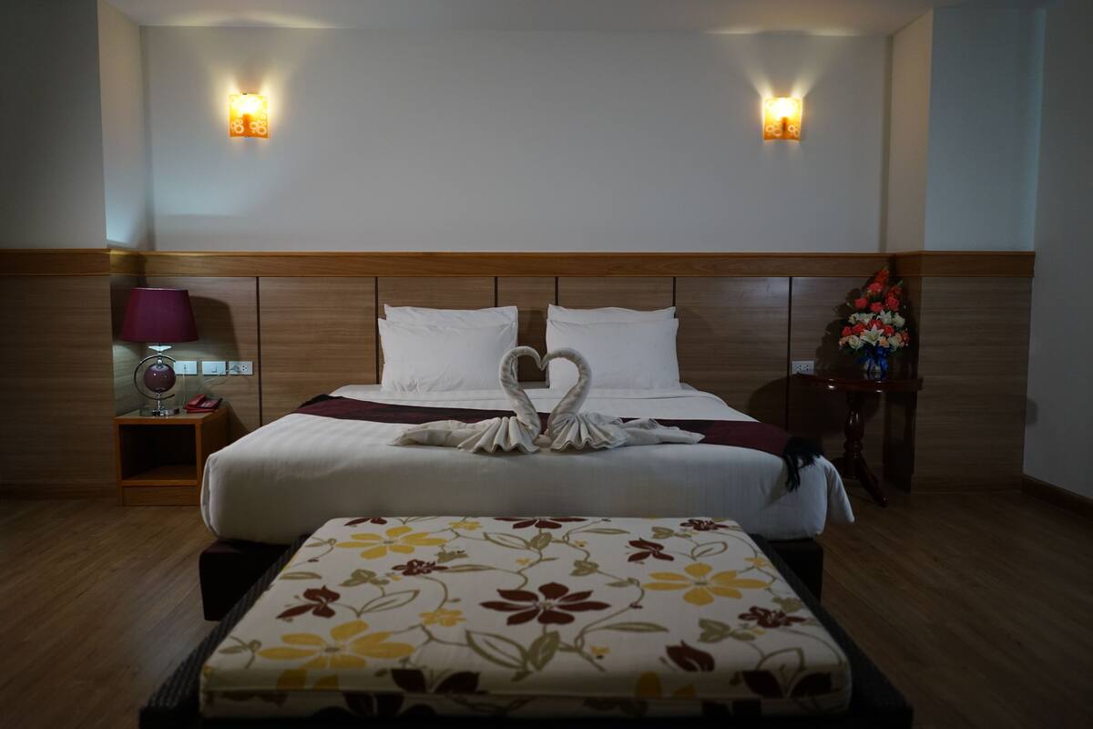 A hotel room with a wooden headboard, a bed with white linens, a decorative swan pillow, a floral-patterned ottoman, and two wall-mounted light fixtures.