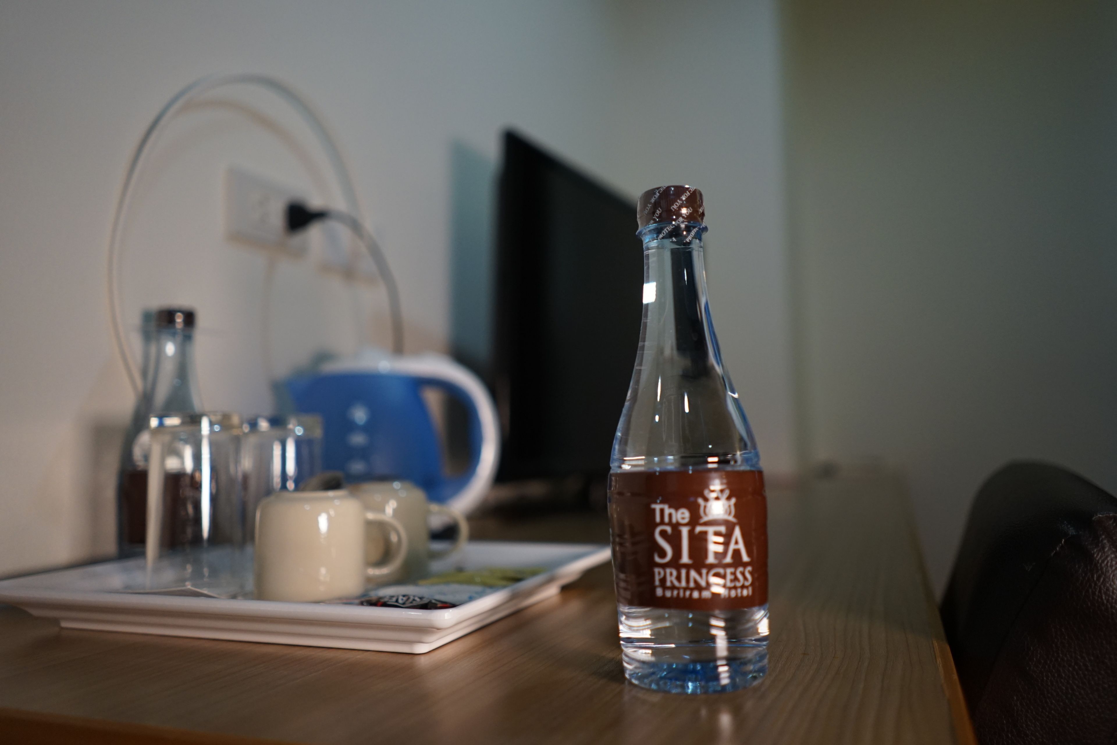 Une bouteille d’eau de la marque The Sifa Princess est posée sur une table en bois, avec une tasse blanche et une tasse bleue en arrière-plan.