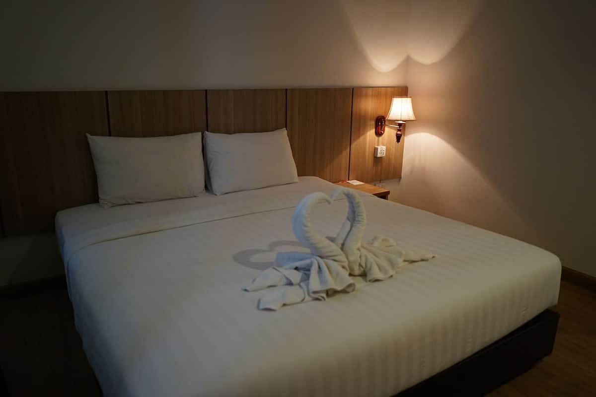 A hotel room with a neatly made bed, a wooden headboard, a wall-mounted lamp, and a bedside table with a lamp.