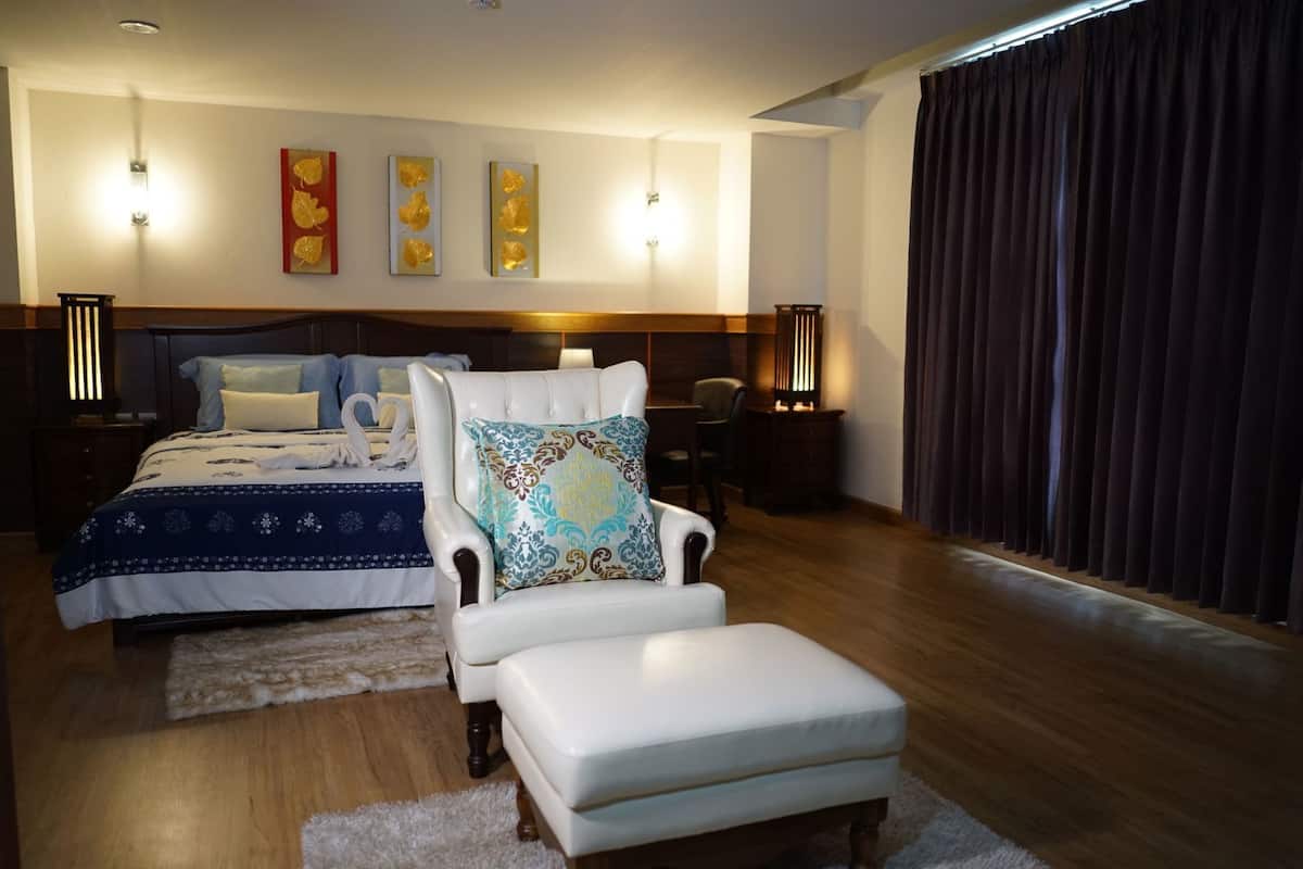 A bedroom with a large bed, a white armchair, a small table, and a large window with curtains.
