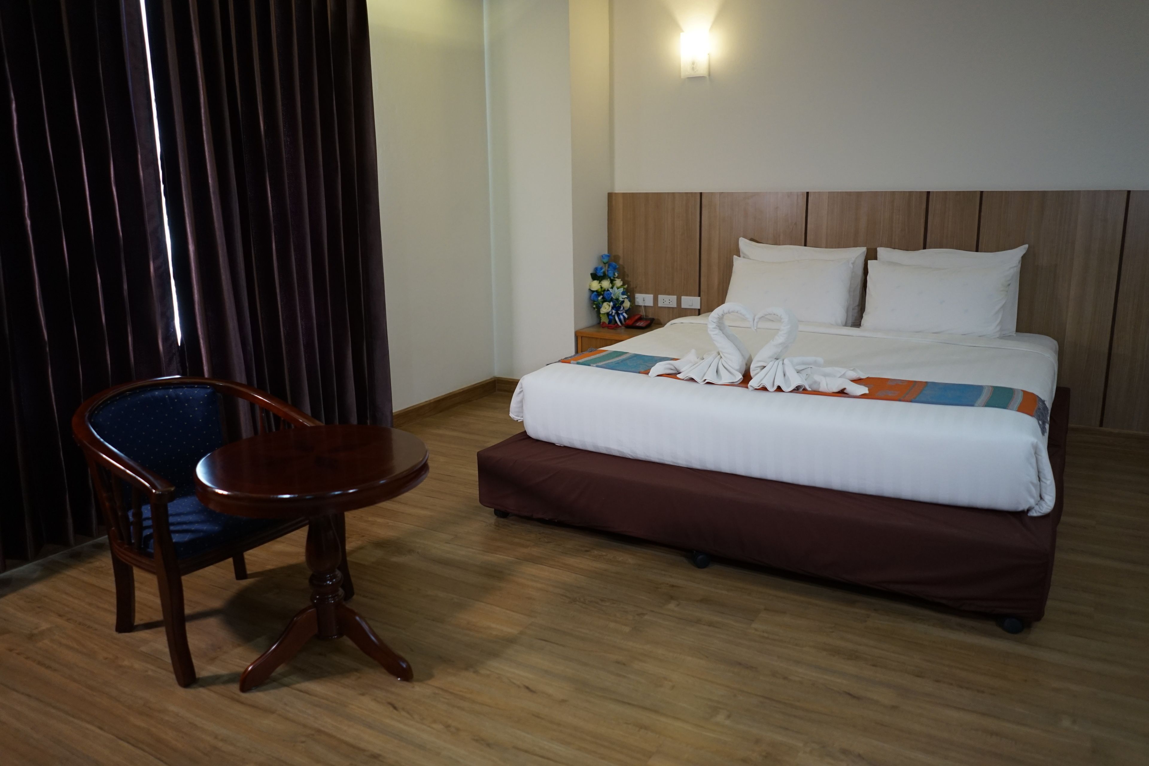 Una habitación de hotel con piso de madera, una cama con ropa de cama blanca y marrón, una mesita de noche de madera con un jarrón de flores y una silla de madera.
