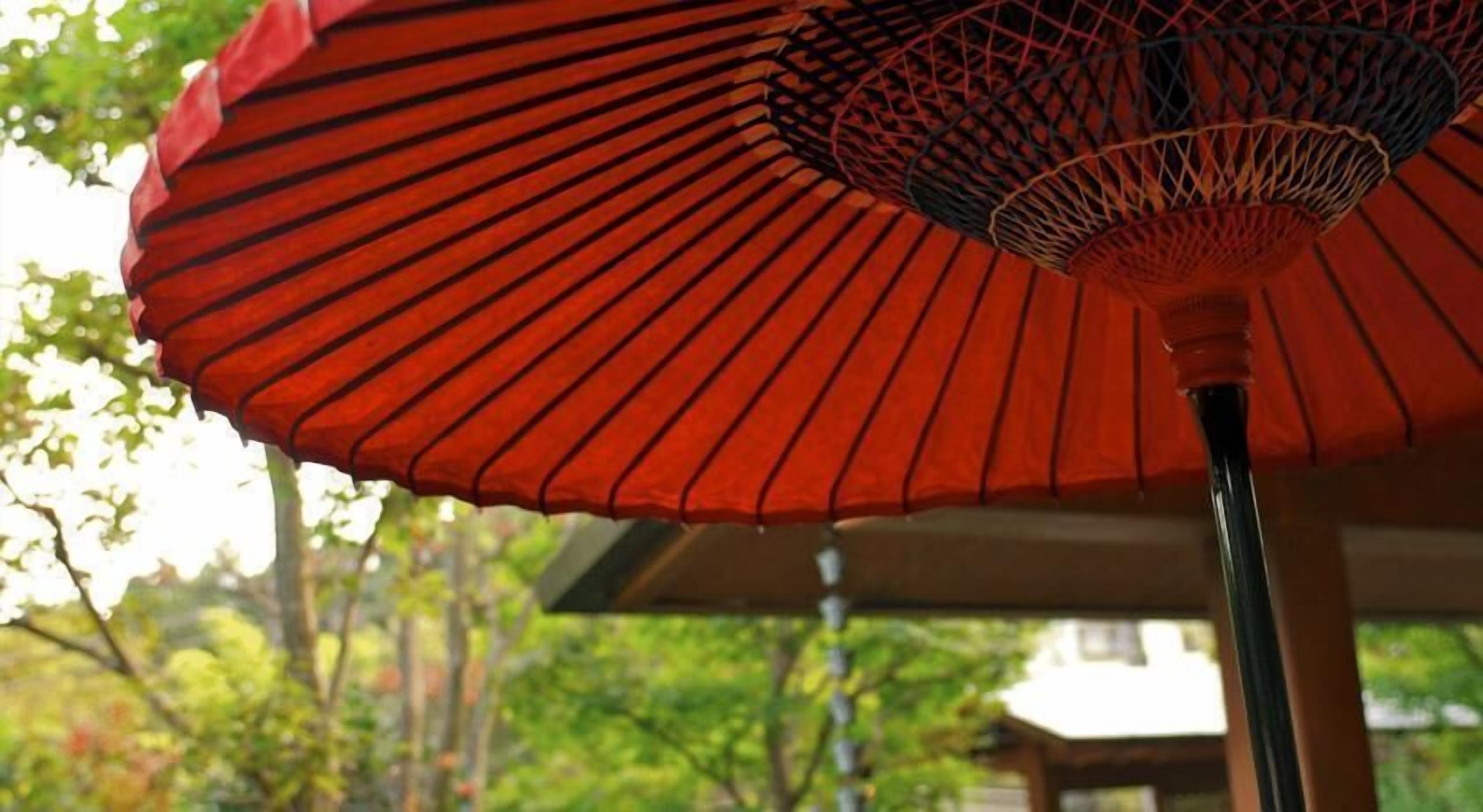 A red umbrella with a patterned design, likely a parasol, in an outdoor setting with trees and a building in the background.
