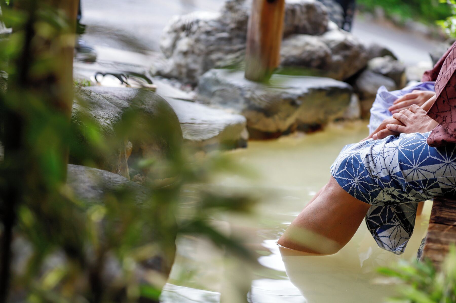 Eine Person sitzt an einem Teich, im Hintergrund befinden sich Felsen und viel Grün.