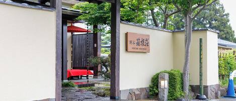Un edificio japonés tradicional con un sendero de piedra que conduce a una entrada cubierta. Se observa un paraguas rojo debajo de la entrada, y una señal con caracteres japoneses está colocada en la pared.