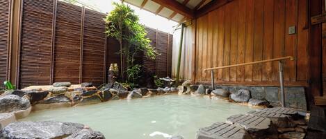 A traditional Japanese onsen with a wooden interior, a stone bath surrounded by rocks, and a bamboo fence in the background.