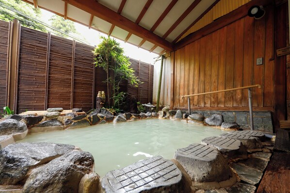 Ein traditionelles japanisches Onsen mit Holzvertäfelung, einer steinernen Badeanlage inmitten von Felsen und einem Bambuszaun im Hintergrund.
