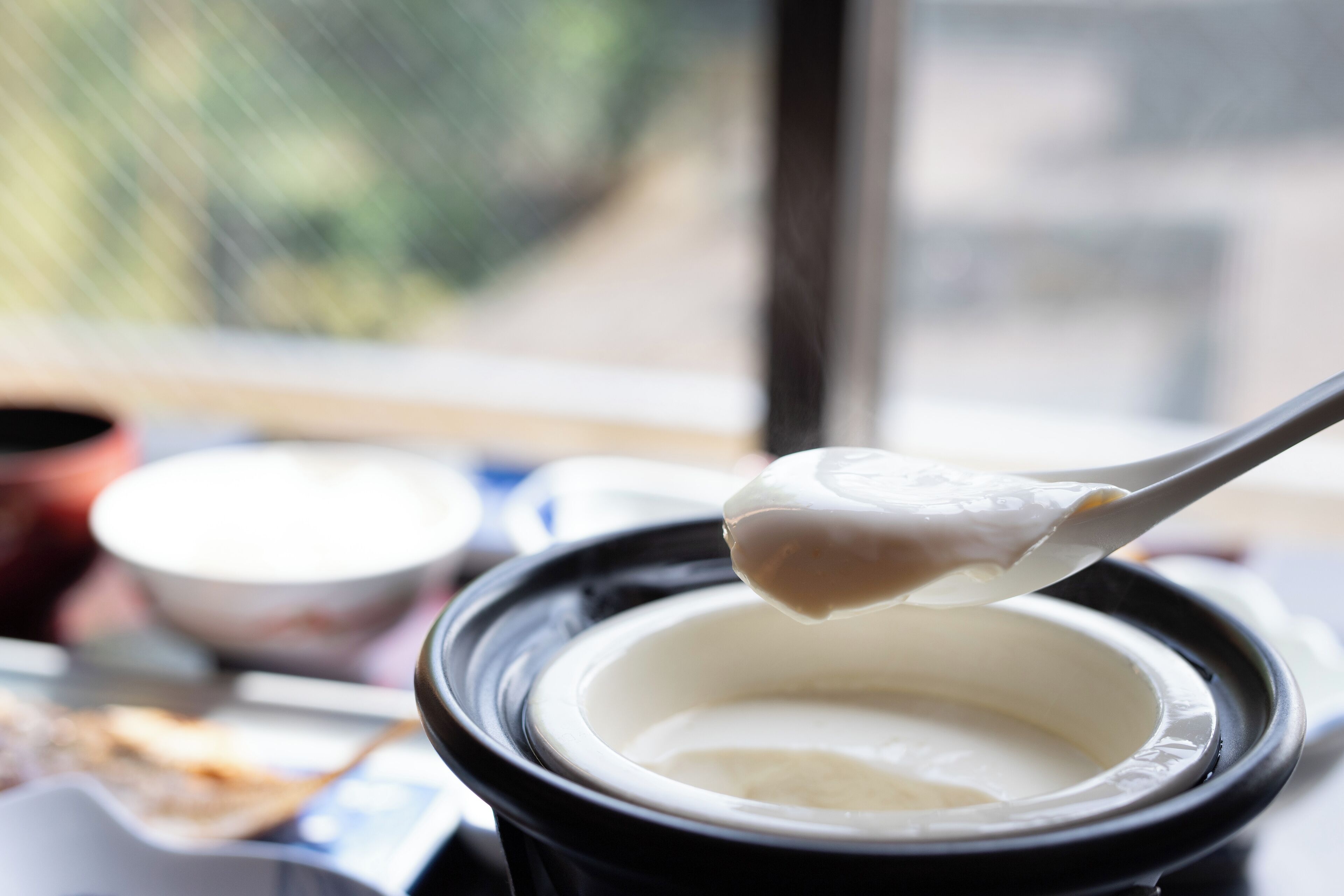 A close-up of a spoon lifting a creamy substance from a black container, with a blurred background suggesting an indoor setting.