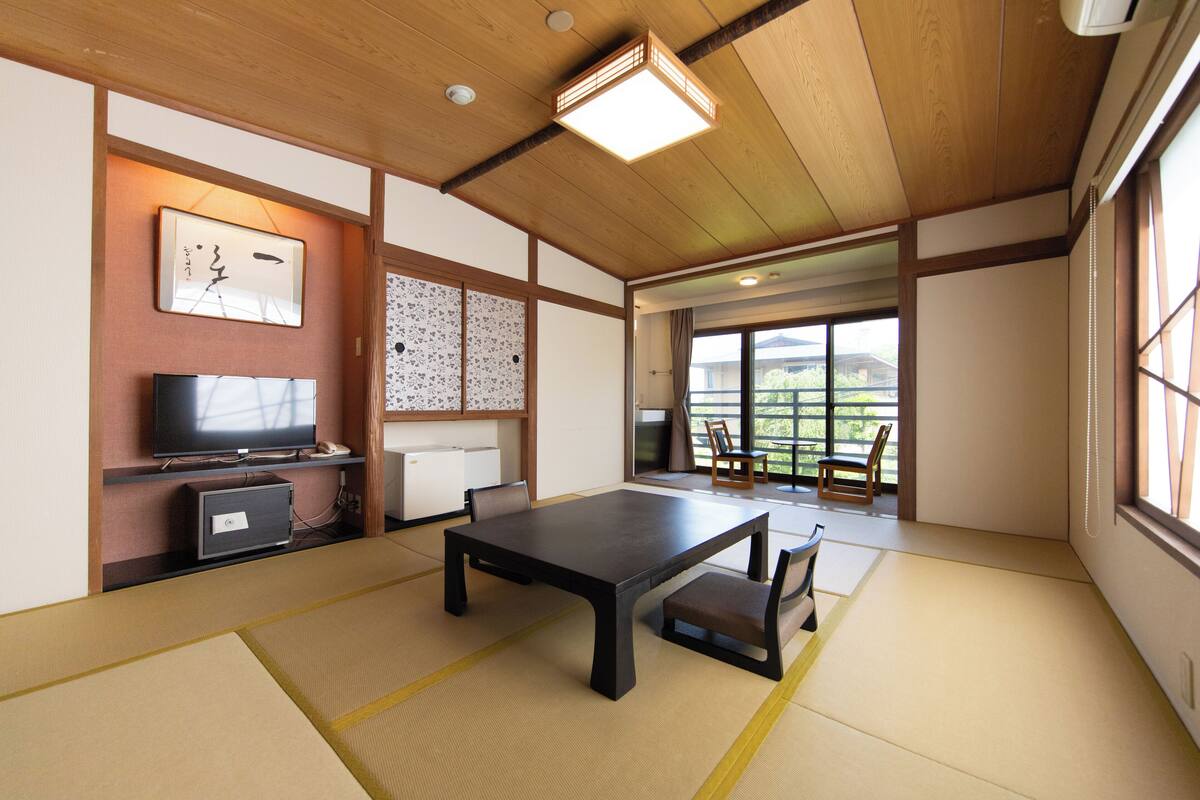 A traditional Japanese room with tatami flooring, a low table with a bench, a television, and sliding doors leading to a balcony with a view of greenery.