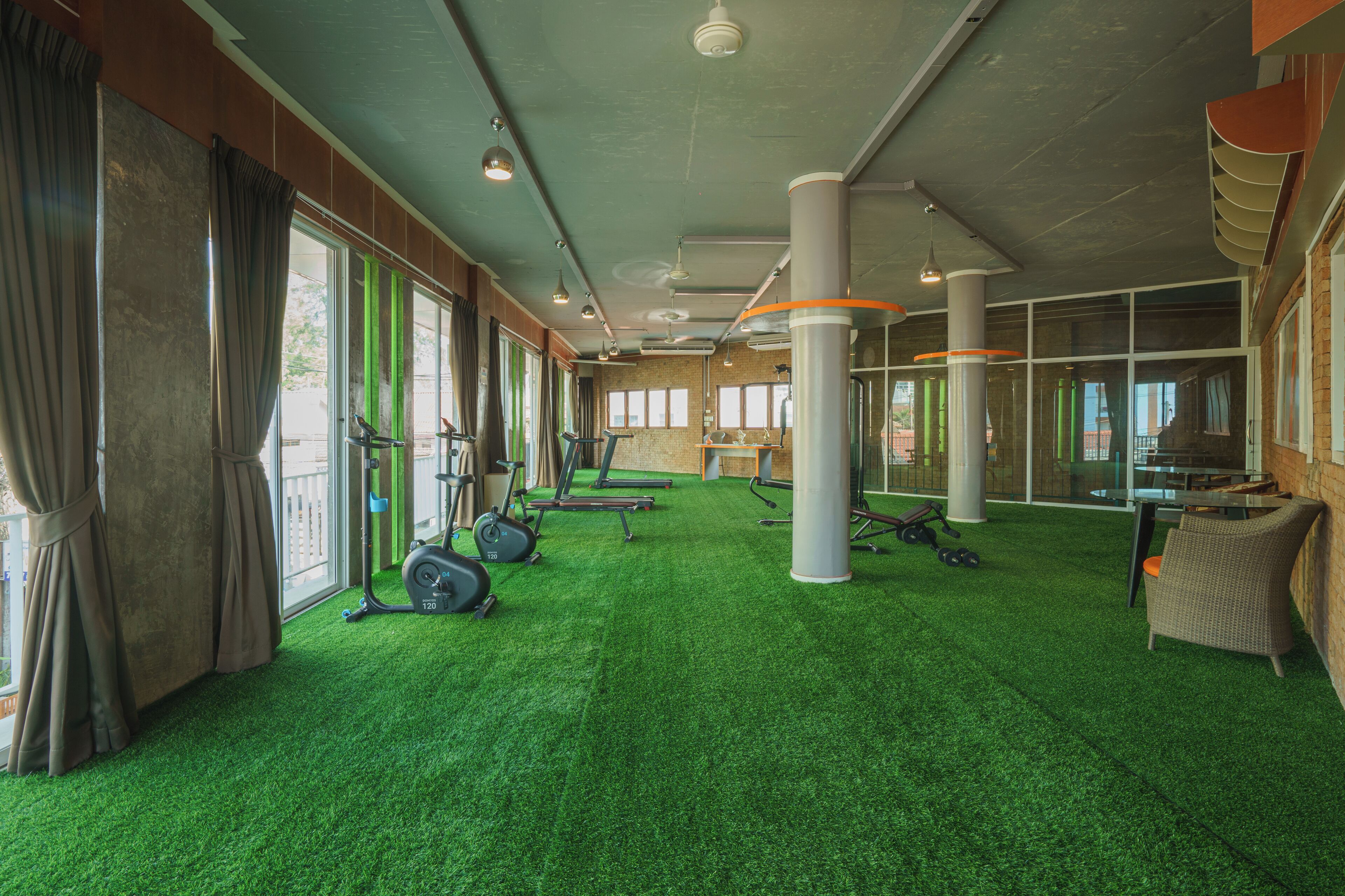 Une salle de sport équipée de matériel d’exercice, dotée d’un revêtement de sol en gazon synthétique vert, de grandes fenêtres et d’un fauteuil en osier.