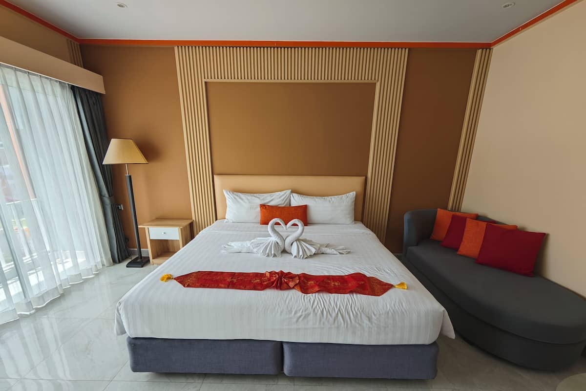 A hotel room with a large bed, a sofa, a nightstand, a lamp, and a view of the outdoors through sheer curtains.