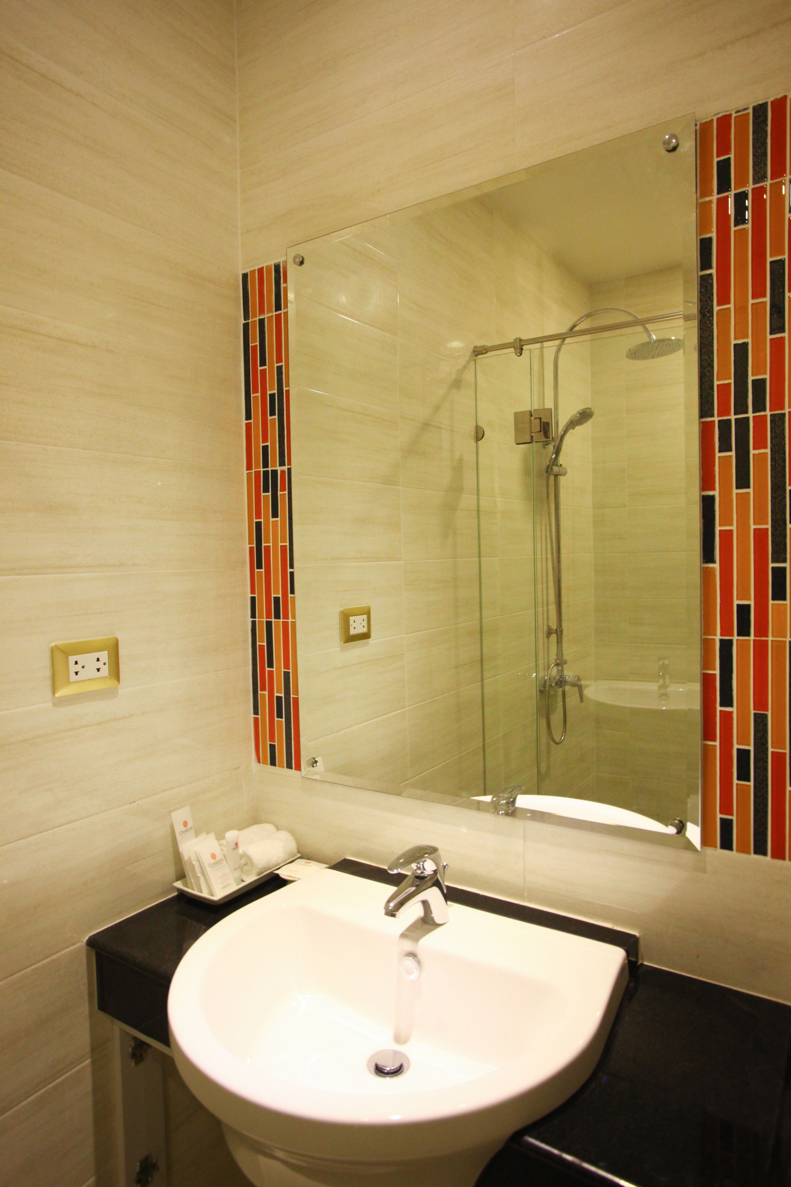 Un baño con un lavamanos blanco, cabina de ducha con mampara de vidrio y espejo con un borde de azulejos de colores.