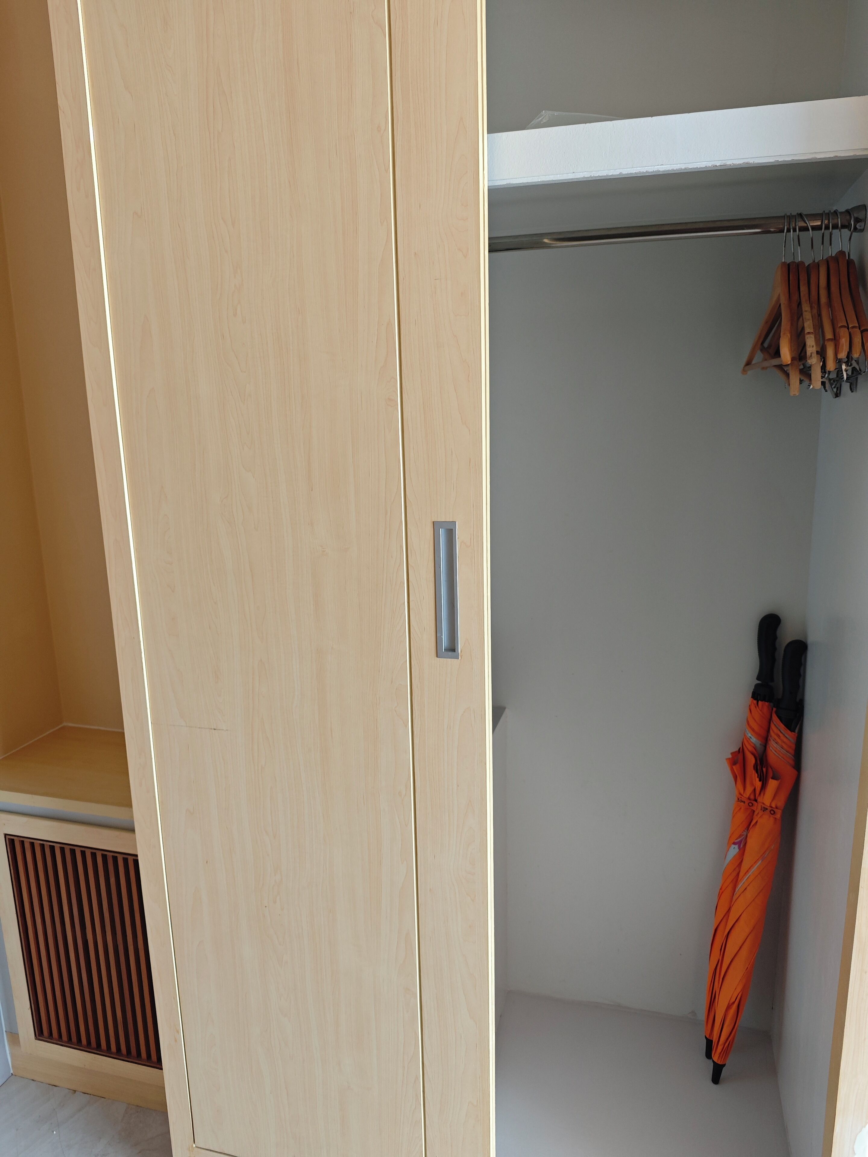 A wooden wardrobe with a handle, a vent at the bottom, and an orange umbrella hanging on a rod.