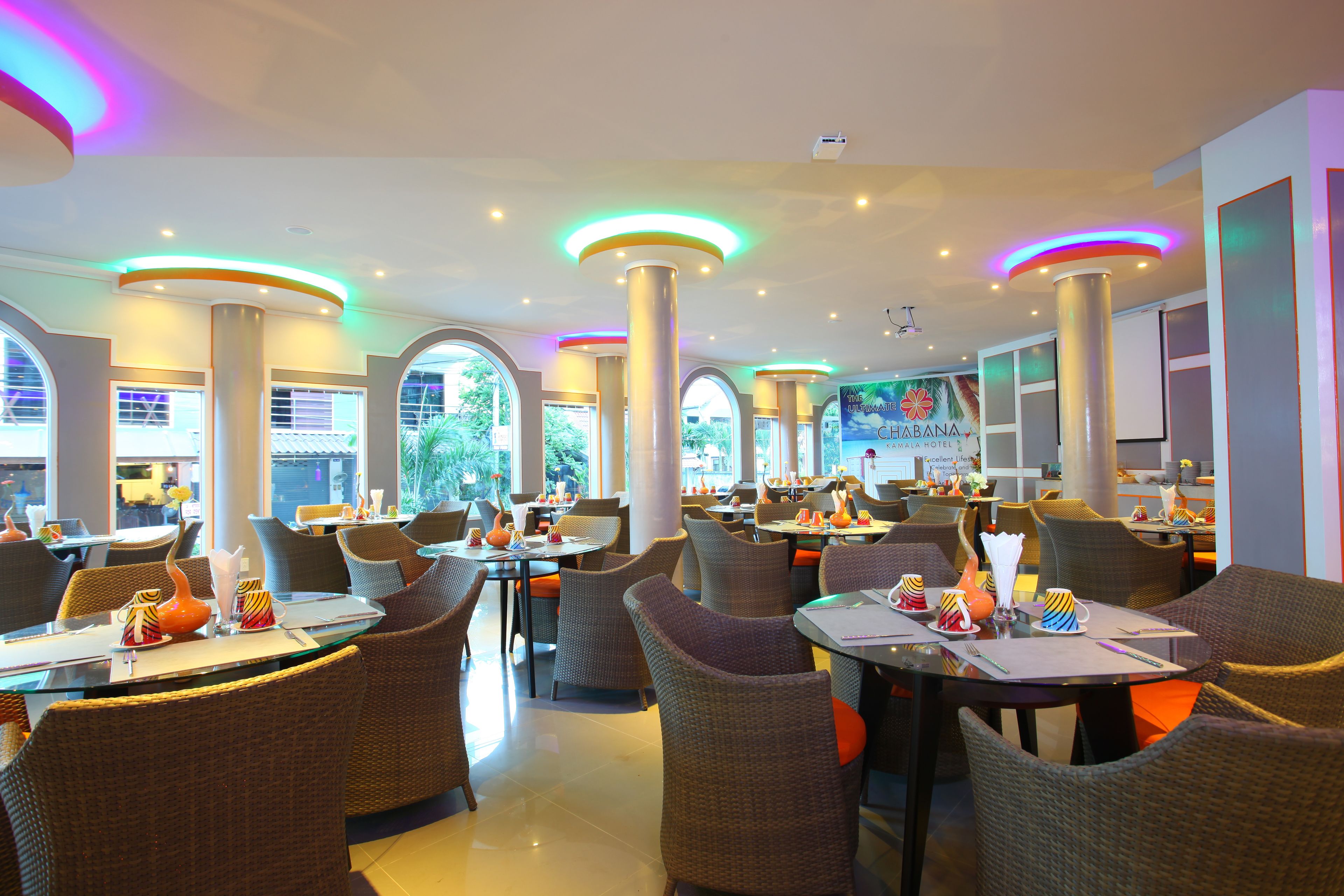 Un restaurante con mesas altas y sillas de mimbre, ventanas amplias y lámparas de techo coloridas.