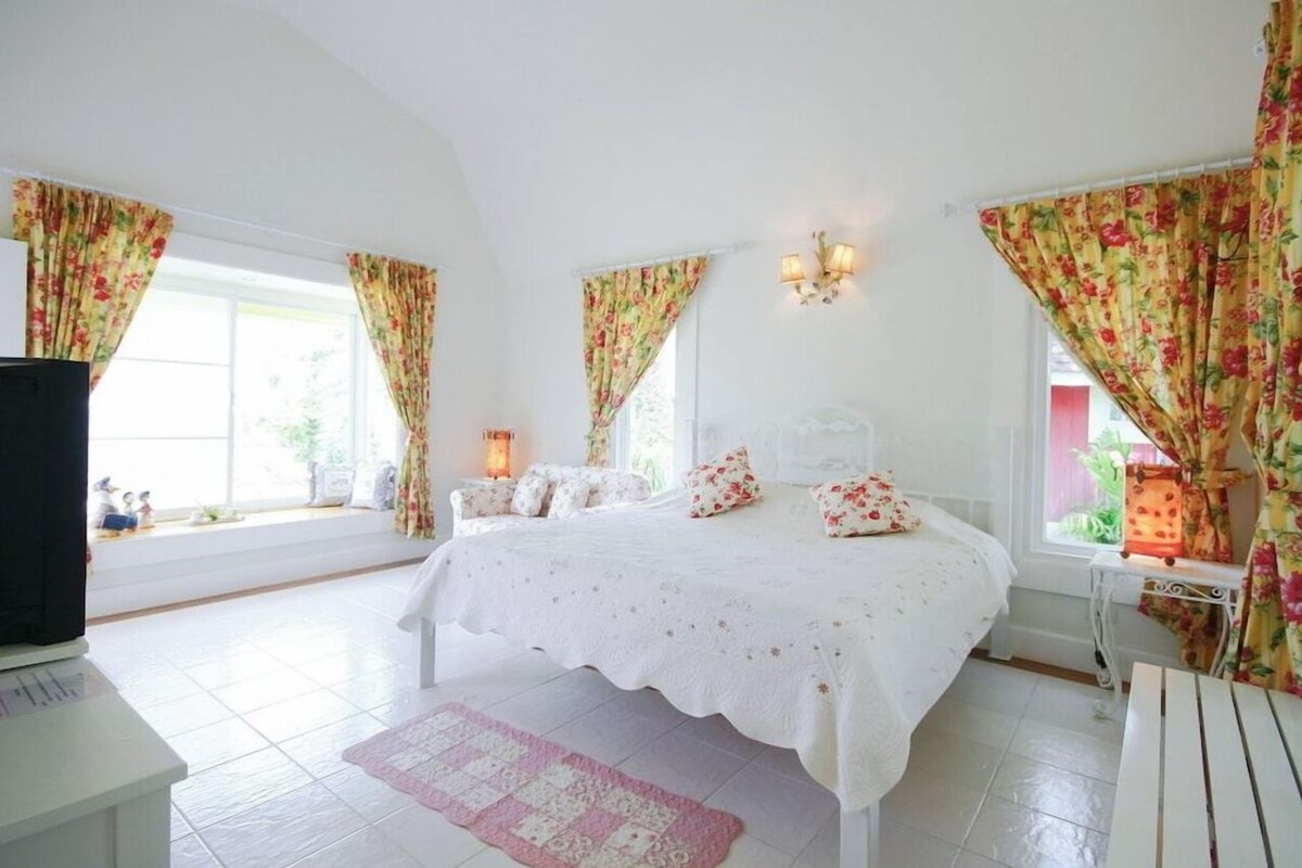 Een slaapkamer met een bed, gordijnen, een nachtkastje, een lamp en een raam.