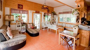A cozy living room with a fireplace, wooden beams, a checkered table, and a window with a view of greenery. - Belmont Village (Pak Chong)
