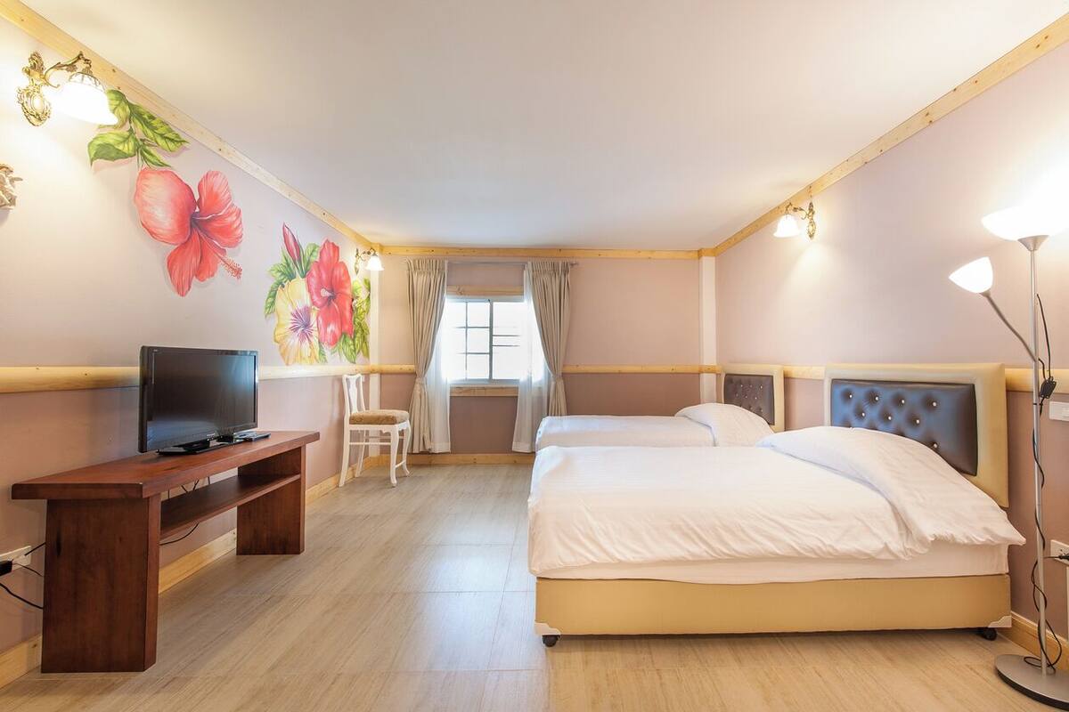 A hotel room with a wooden floor, a TV on a wooden stand, two beds with white bedding, a desk, and a wall with floral artwork.