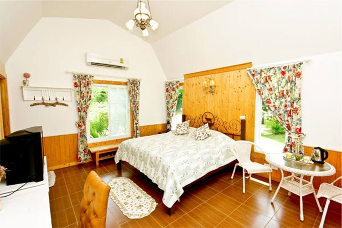 A bedroom with a bed, a desk with a chair, a small table with a chair, an air conditioning unit, and a window with curtains.