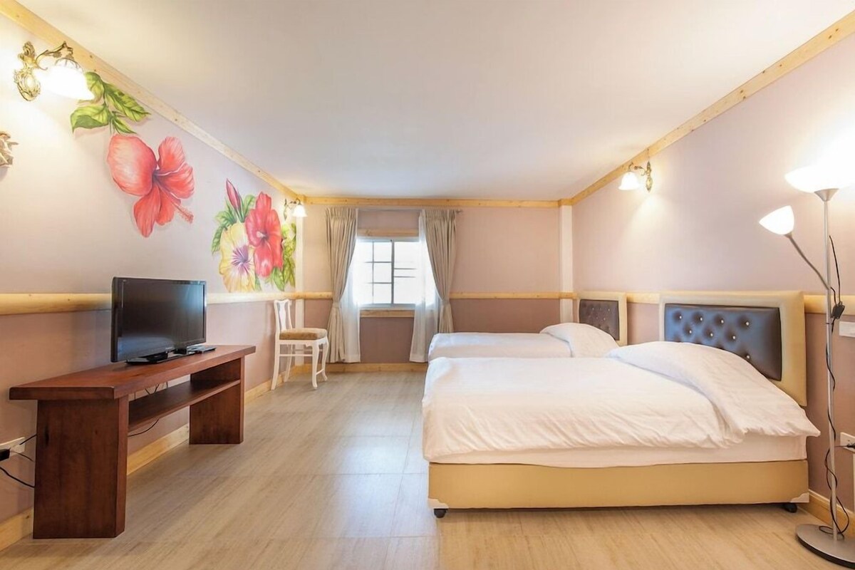 Een hotelkamer met een houten vloer, een televisie op een houten standaard, twee bedden met witte beddengoed, een bureau en een wand met bloemenkunst.