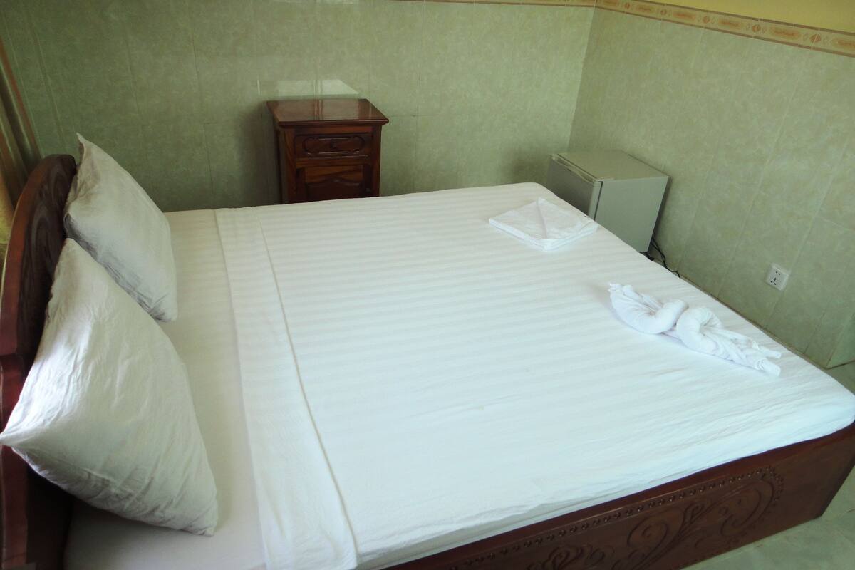 A neatly made bed with white linens and a folded towel, a wooden nightstand with a drawer, and a small bedside table with a lamp.